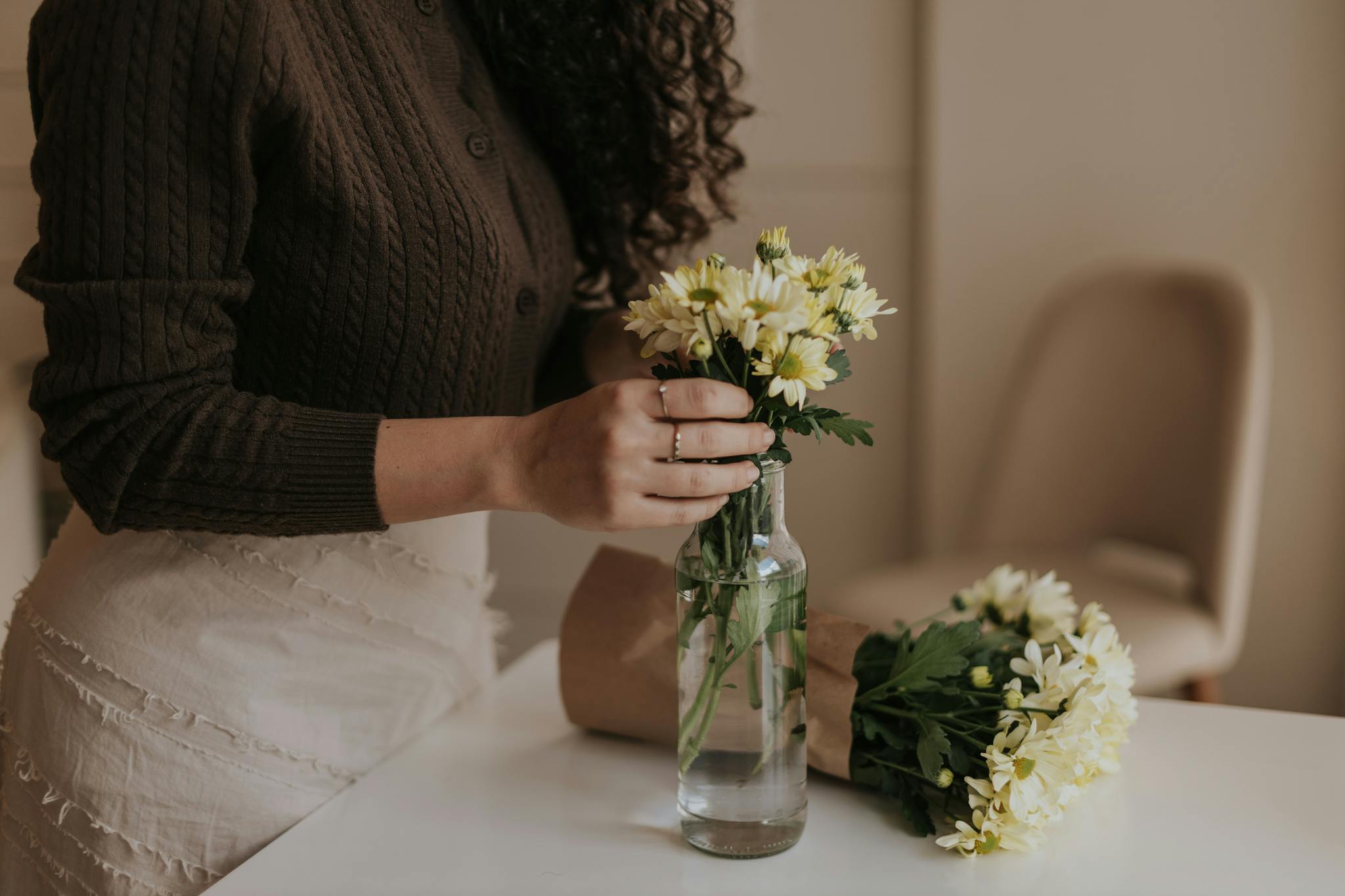 A woman in a cozy sweater arranges yellow daisies in a glass vase indoors, creating a warm atmosphere.